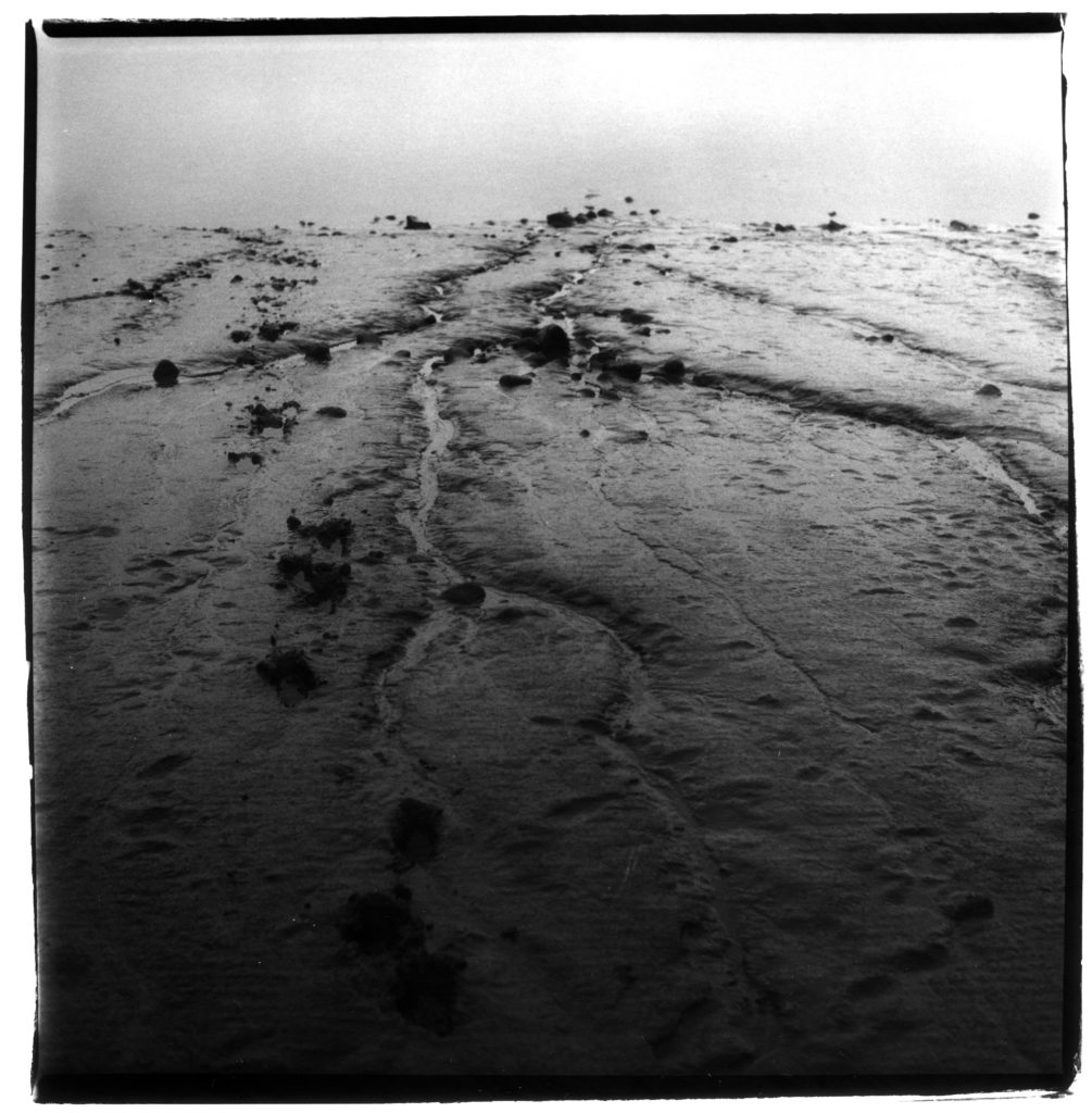 A black and white photograph of sand, showing patterns made by rivulets. The image goes from dark at the bottom to bright white at the top.
