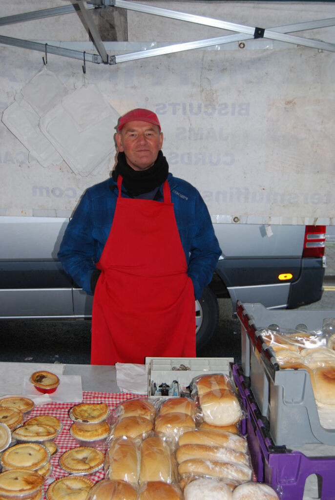 A colour photography of a male stall holder wearing a red apron stood in front of a display of baked goods on a market stall.
