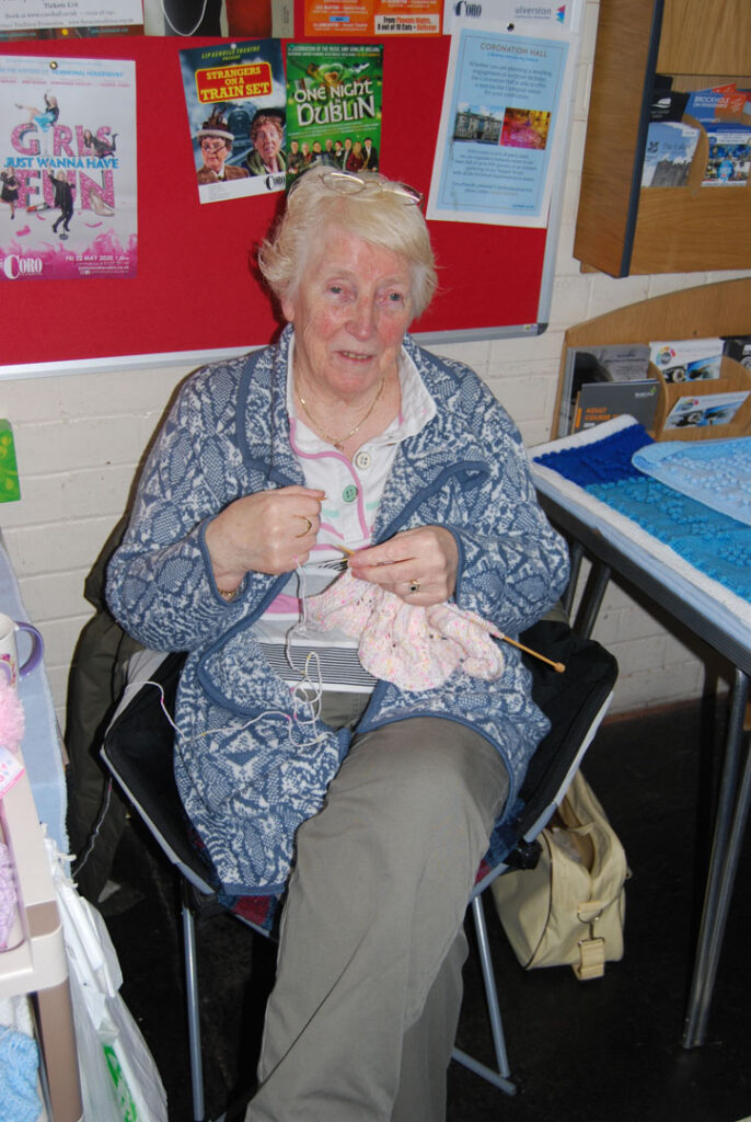 A colour portrait photograph of a woman sitting down knitting wearing a blue jacket.