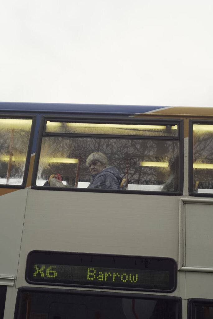 A colour photograph of a close up of a person on the top run of a double decker bus. The sign for the bus says it is the X6 Barrow.