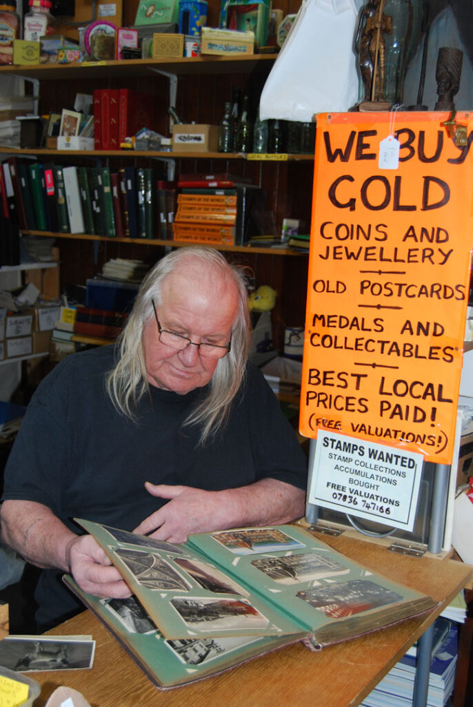 A colour portrait of a man with long white hair sitting down looking through a book of old photographs in an album. Behind him is a bright orange sign that says "We buy gold coins and jewellery old postcards medals and collectibles. Best local prices paid!