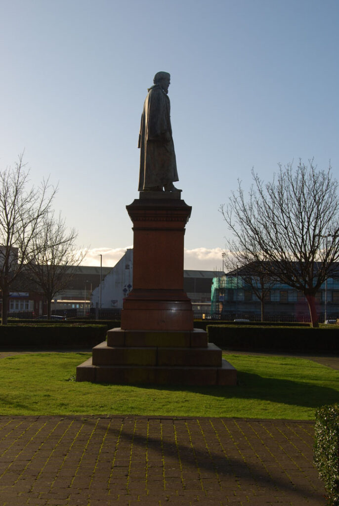 A photograph of a stone statue on a plinth in the middle of a public garden. The sky is blue and the trees in the background are bare of leaves. The photograph is taken by the side.