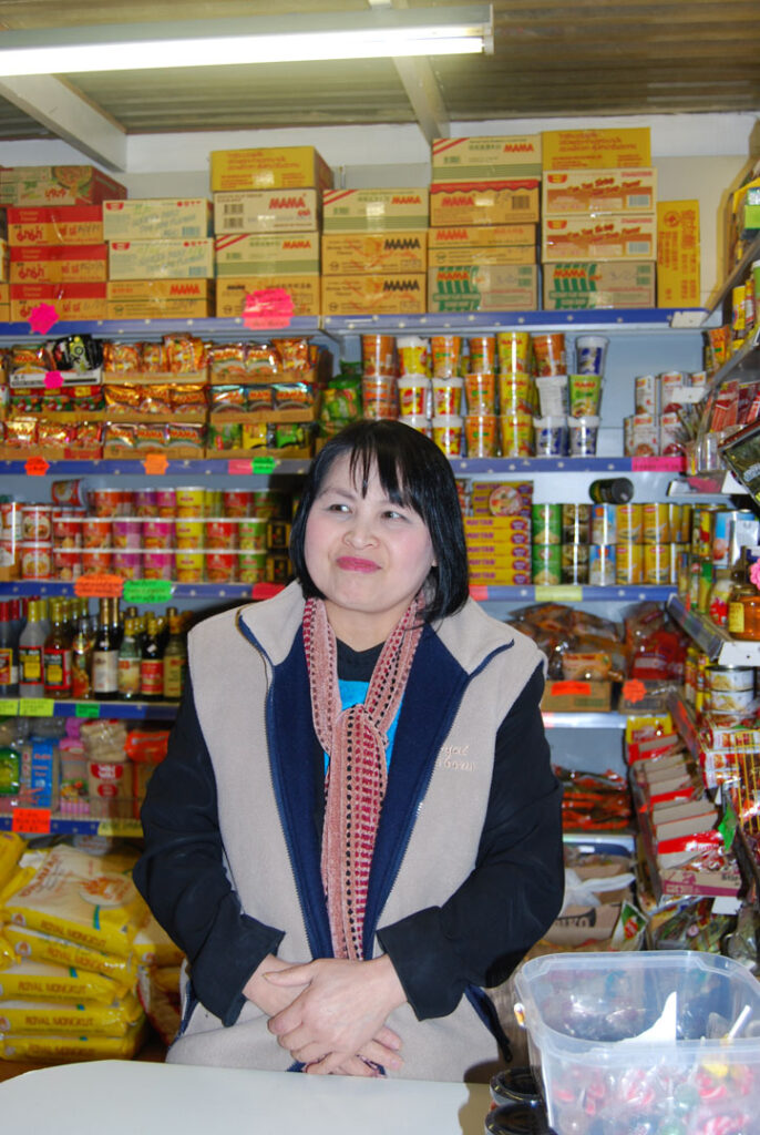 A colour portrait photograph of a women with dark hair sat behind a till in a shop.
