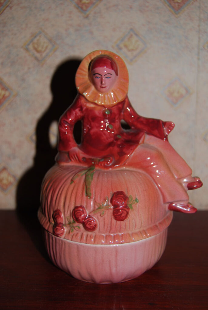 A still life colour photograph of a ceramic bowl with a lid which has a sculptural person sat on top in a red outfit.