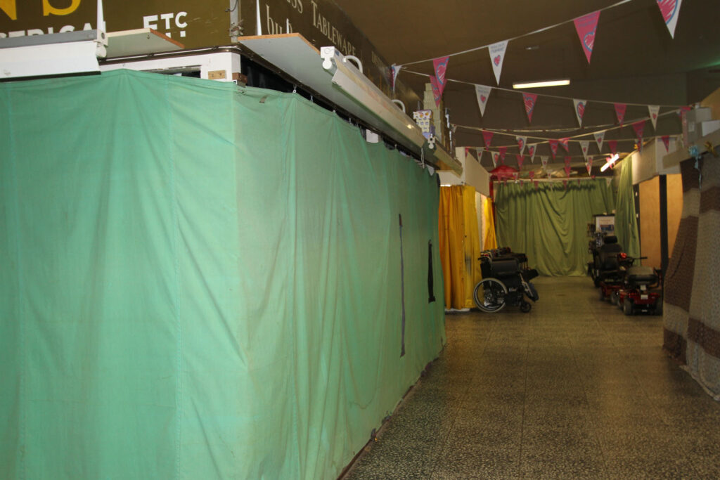 A colour photograph of a market stall in an indoor market with a green cover over the sides of the market stall showing it as closed. In the distance there is a collection of wheelchairs and mobility scooters. Pink and white bunting hangs from the ceiling.