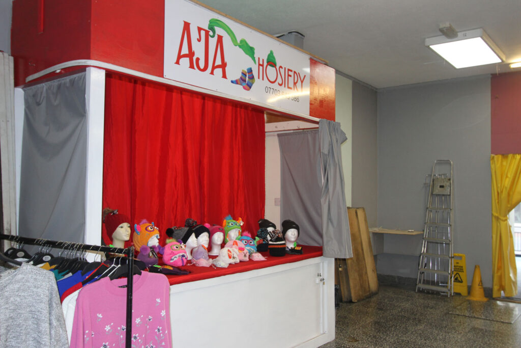 A colour photograph of a market stall in an indoor market. The stall is called AJA Hosiery with a red and white sign. On the market stall there are a range of model heads with different hats on top. In the foreground of the photograph are some tshirts on hangers on a rail.