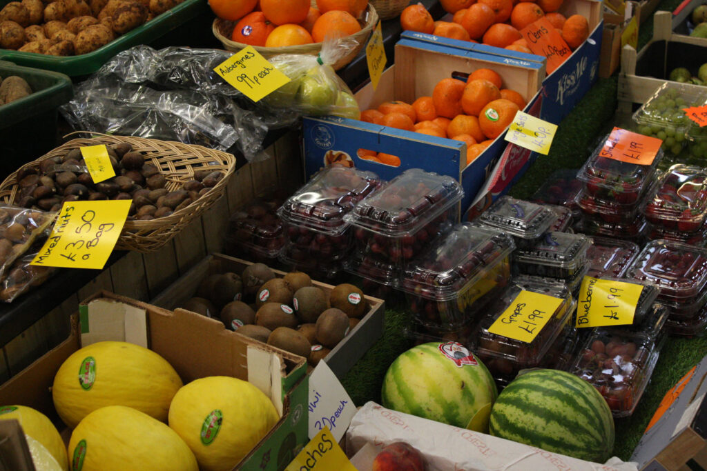 A close up colour photograph of fruit on a market stall. The fruits are in boxes with yellow handwritten signs displaying their prices. Fruits displayed include mixed fruits, melons, blueberries, grapes, aubergines, strawberries and clementines.