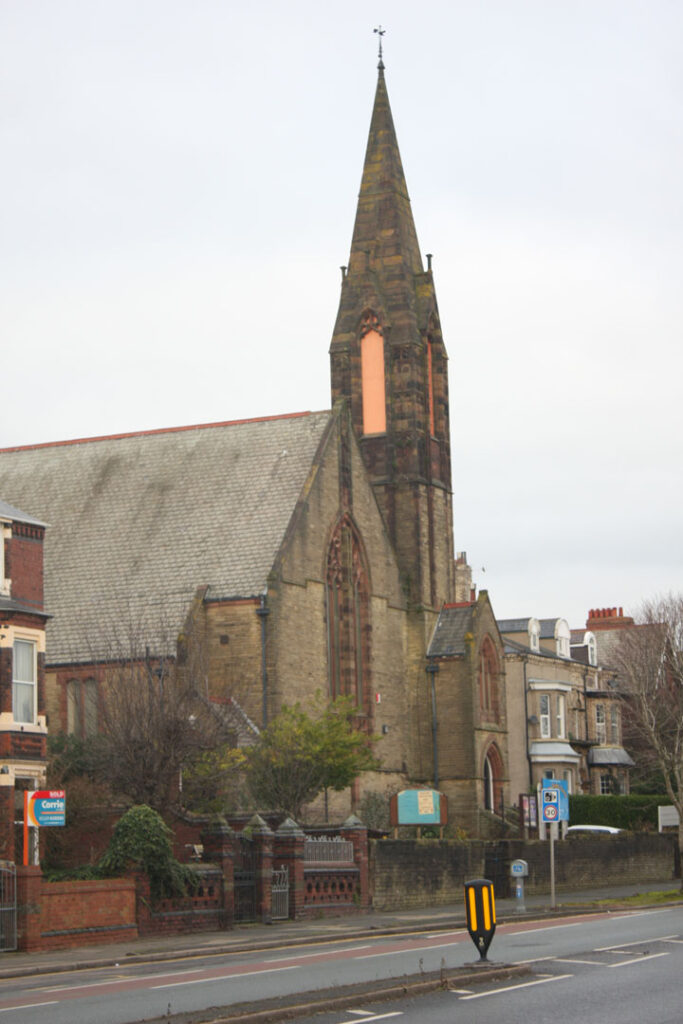 A colour photograph of a church with a spire shot from across on the other side of the road.