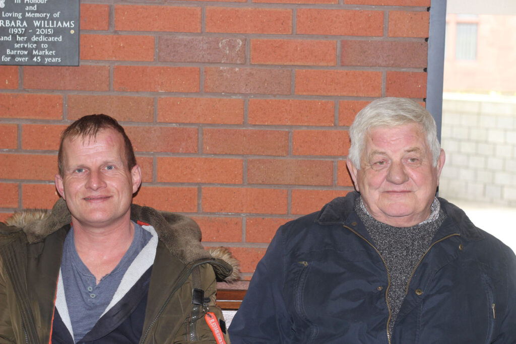 A colour photograph of a portrait of two white men one with brown hair and the second with white hair. They are stood in front of a brick wall.