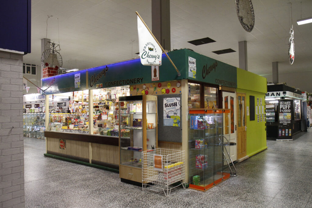 A colour photograph of a market stall called Cherry's confectionery. The market stall is green and is in the middle of an indoor market hall.