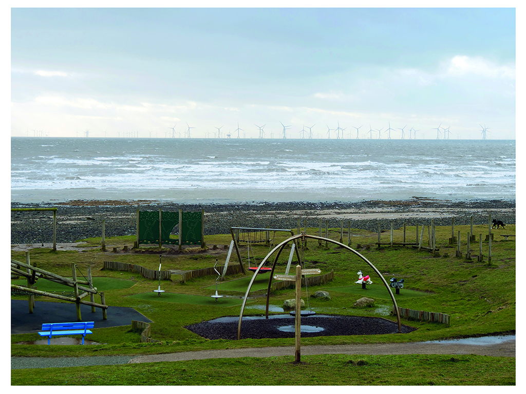 A colour landscape photograph of a kids playground in front of the sea. In the distance is an off shore wind farm. The playground is empty of children apart from a black and white dog in the far right of the picture.