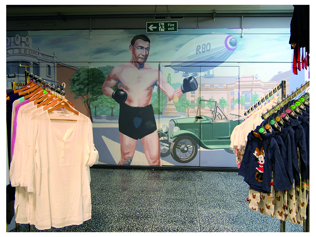 A colour photograph of the interior of a shop. In the foreground are some stands with clothes for sale on hangers. In the background on the far wall there is a handpainted mural of a boxer, a vintage green car and an airship.