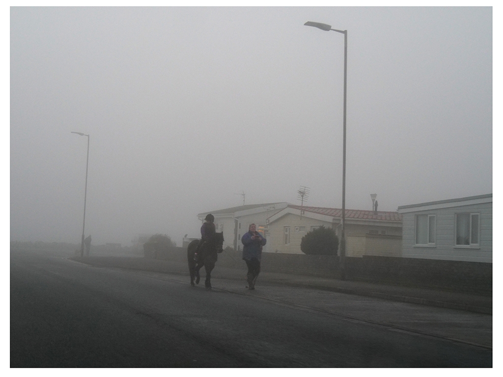 A photograph of a child riding a horse with a person walking next to them. They are walking down the road with bungalows on one side. The weather is misty and grey.