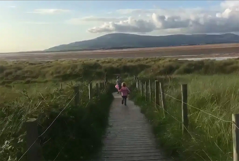 A colour photograph from a video showing two children running towards the beach through grass sand dunes.