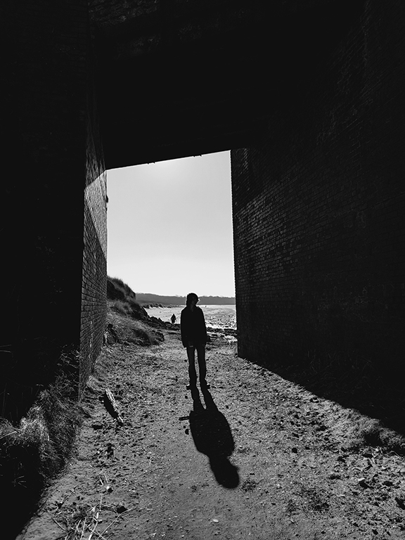 A black and white photograph a person stood in a tunnel. The sun is behind them so they are in silhouette. Through the entrance to the tunnel you can see a glimpse of the coastline.