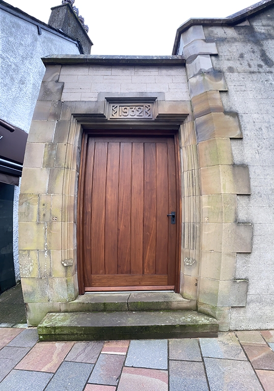 A colour photograph of a wooden door in a stone building. On top of the door is a stone carved sign with the date 1932 on it.