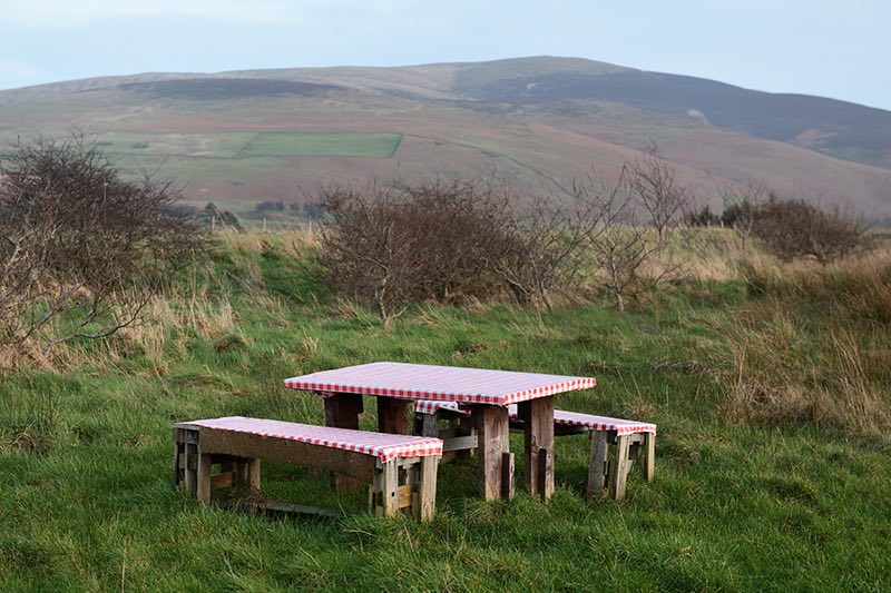 A colour photograph with a picnic bench in the foreground of a rural landscape. The wooden picnic bench has a red and white gingam cloth over the top. In the background there is a hill top.