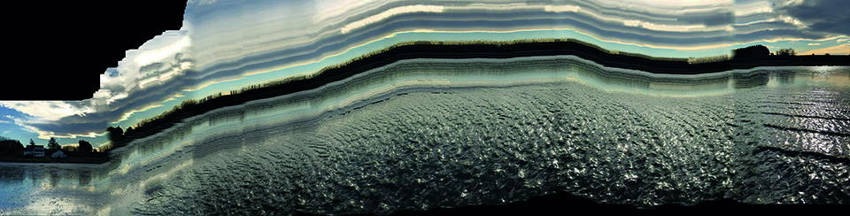 A colour photograph of water with ripples on and in the horizon is the edge of the water with a small building and trees in black silhoutte. The sky has clouds with the sun behind them. The photograph has been stretched in to a panoramic containing multiple photographs in one stitched together.
