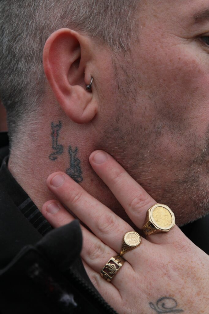 A colour close up photograph of a man taken by the side. The man has two tattoos underneath his right ear. His hand is held up to his face and has three rings on three of his fingers. His ear has a ring earring in it.