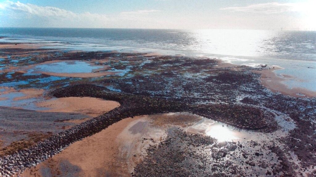 A colour landscape photograph of a beach on the west coast of cumbria. On the beach there are various rockpools and a pile of rocks in the shape of an anchor.