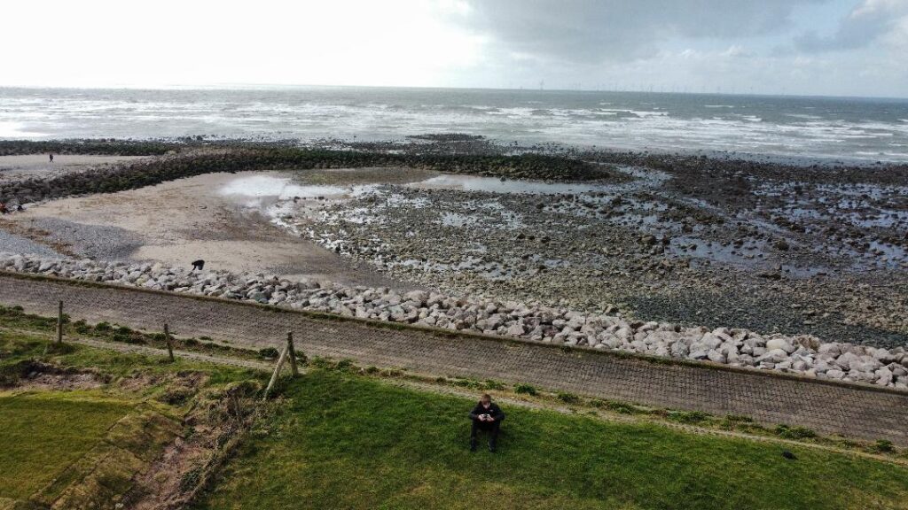 A colour landscape photograph of a boy sat down on a patch of grass by the sea. With the sea in the background with off shore wind farm in the far distance.