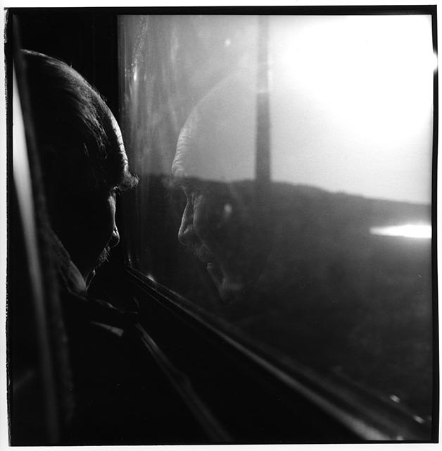 A black and white photograph of a man, with grey hair and a moustache, looking out of a train window. His reflection is visible in the windows glass.