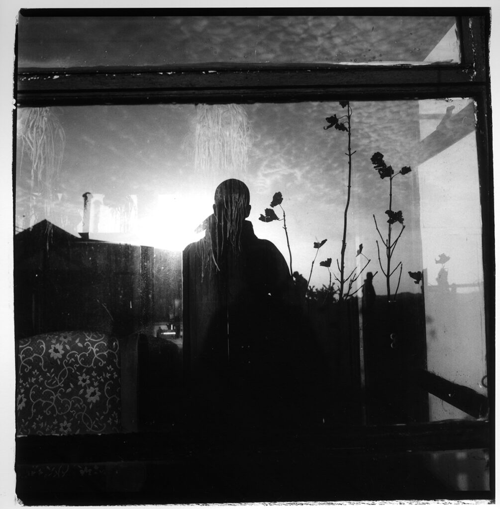 A black and white photograph showing the silhouette of a person reflected in a window. In the foreground there is a floral armchair. In the background there is the outline of a building and some tall plants.