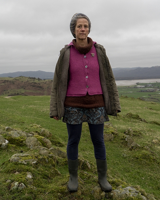 A portrait of a woman stood in a rural setting. The woman is staring in to the camera and is wearing wellington boots and a bright pink cardigan.