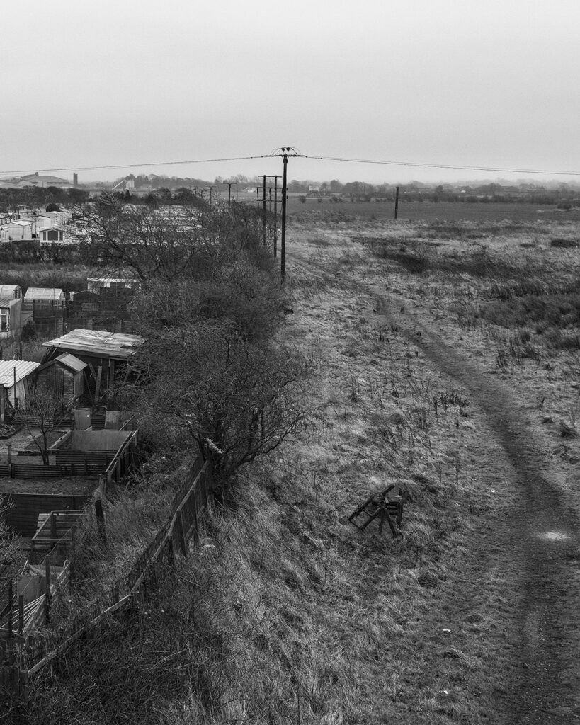 Black and white photo showing the boundary of an allotment. In the foreground there is a broken wooden chair. In the background there is electricity pylons.