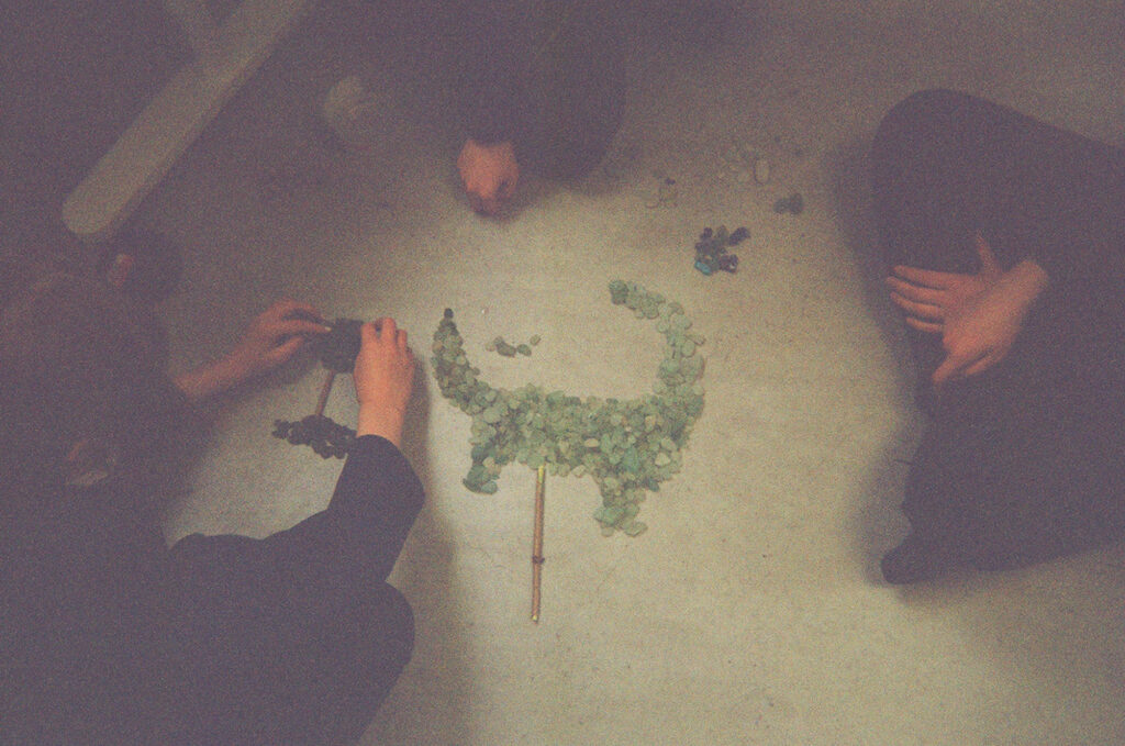 A colour photograph of three people sat around a display of green stones with pencils. The three people are sat cross legged and the camera has taken the photograph from the top looking birds eyes view down to the three people.