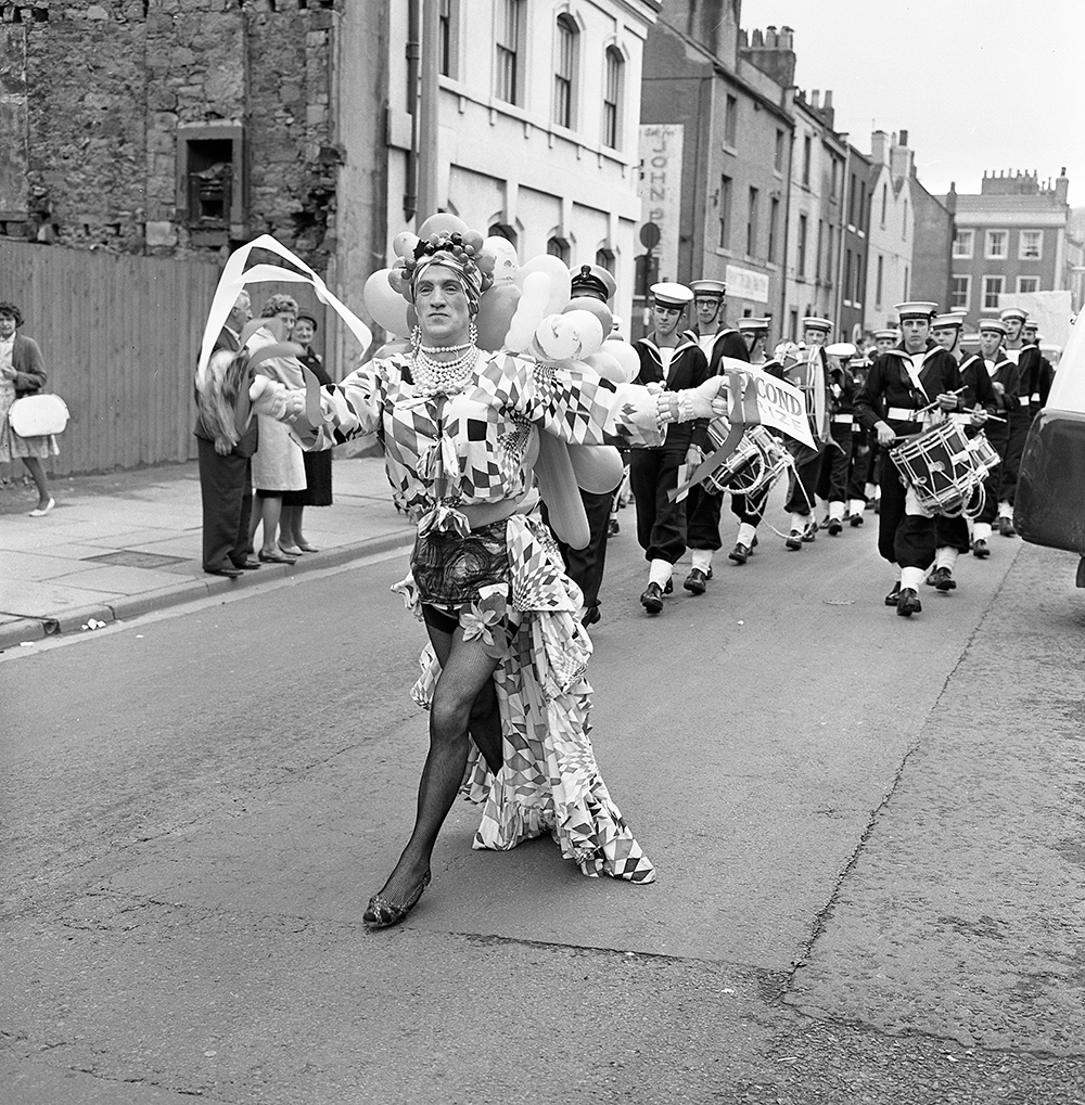 Black and white photograph of a man dressed in drag, walking down a high street. Behind him is a brass band lined up in uniform.