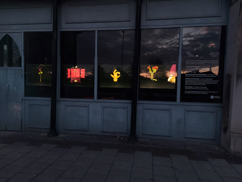 A colour photograph showing four backlit images, displayed horizontally on windows. The images show still life photographs: a rose, a teddy bar and a red jewellery box .