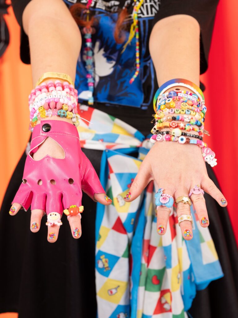Photograph by Casey Orr. Photo is off a pair of hands, outstretched palm down, adorned with colourful jewellery and one pink fingerless glove.